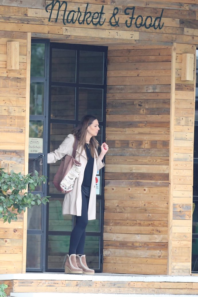 Eva González muy guapa con leggins y jersey en negro con una gabardina en camel, botines de cuña de esparto y un bolso boho en rosa y nude