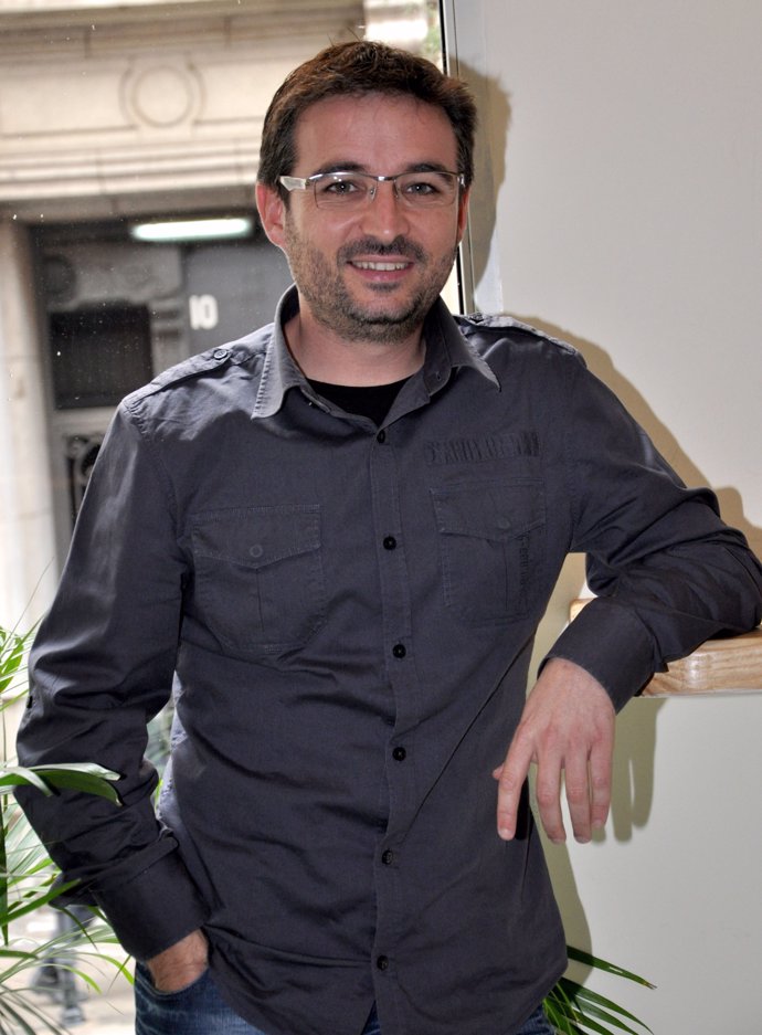 Jordi Évole posando apoyando un brazo en la pared, con camisa gris de sport, pantalón negro, y camiseta negra