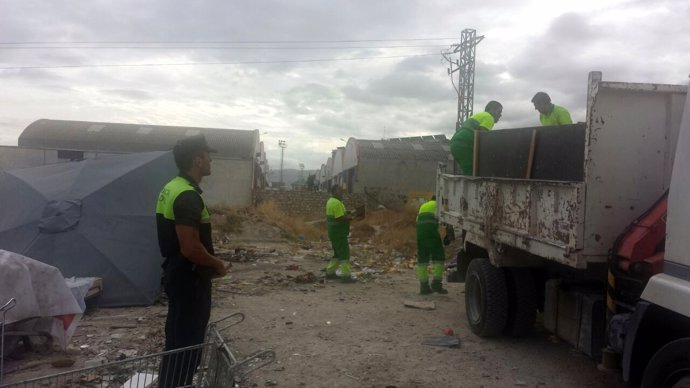 Operarios municipales desmantelan el asentamiento chabolista ante un agente.