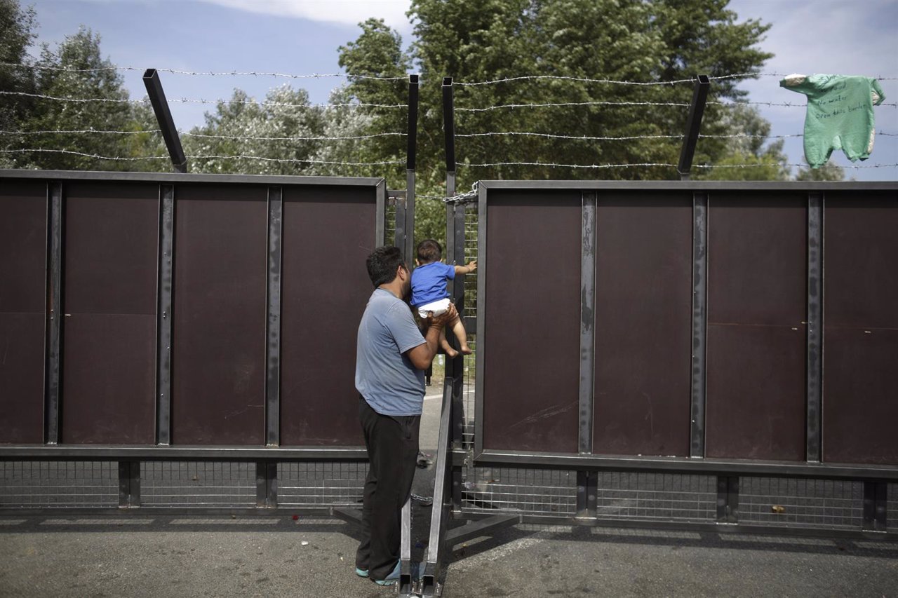 A migrant lifts a child to let it peek on the other side of a barrier at the bor