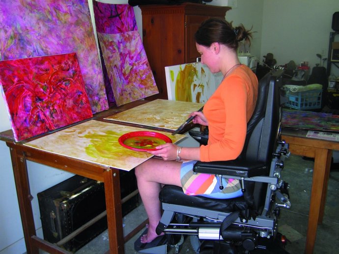 Artist (female wheelchair user) puts paint and brush to canvas in her studio.