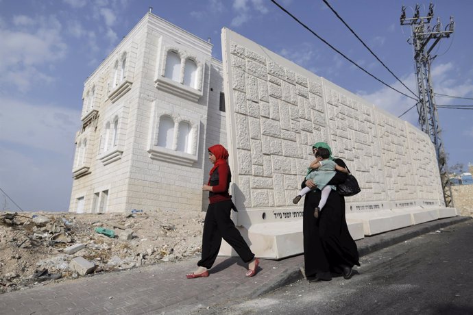Women walk past a newly erected temporary concrete wall that measures around 10 