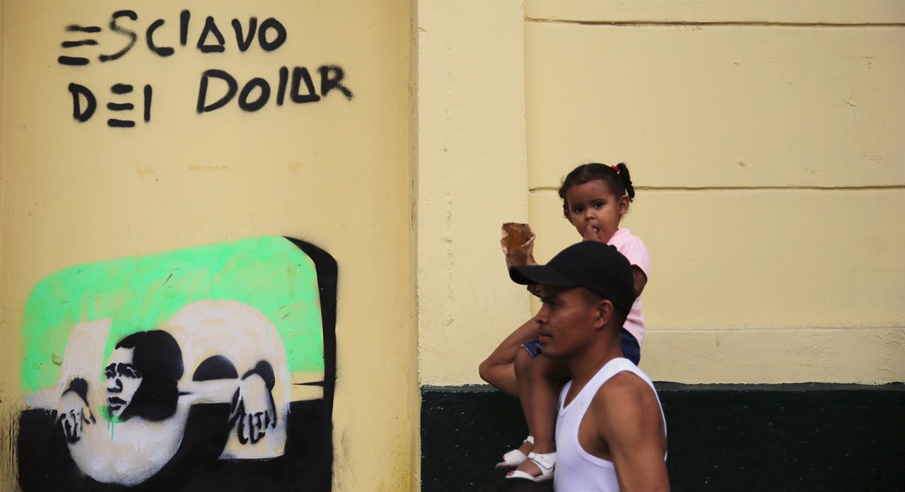 Man carries her daughter as they walk past a graffiti that reads 
