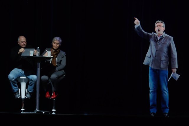 People sit at a table on stage and look towards the hologram of politician Jean-