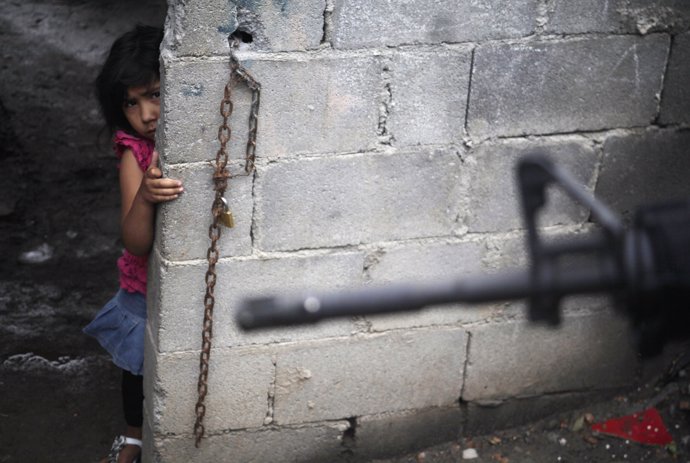 A girl peeks from behind a wall as a police officer patrols a neighborhood in Gu