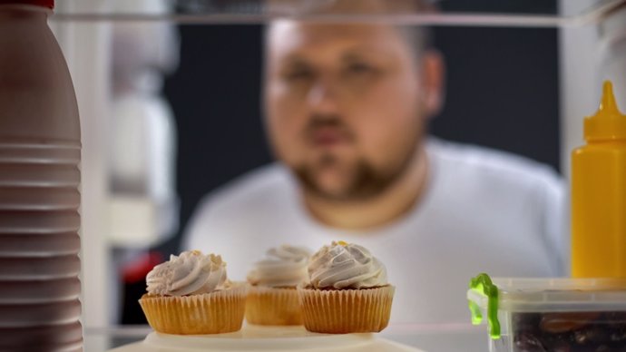 Hombre con sobrepeso, hambriento, mirando pasteles de crema edela nevera por la noche.