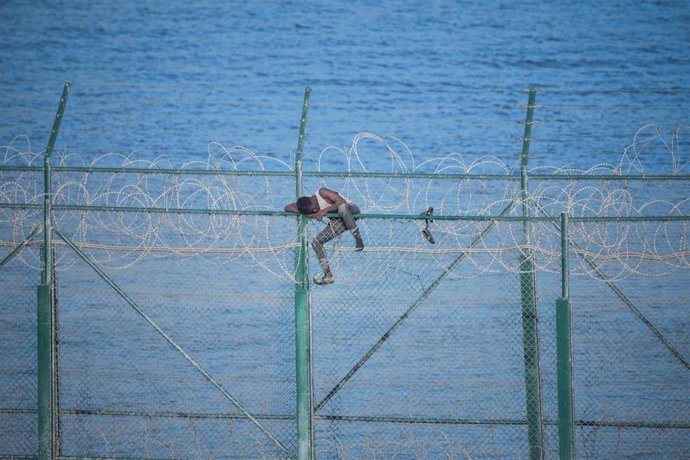 En la imagen, uno de los migrantes que han participado en Ceuta el salto a su doble valla en la primera incursión en grupo en un año se aferra a las concertinas