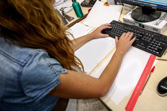 Una dona escriu en el teclat del seu ordinador, amb papers al voltant mentre treballa a l'oficina.