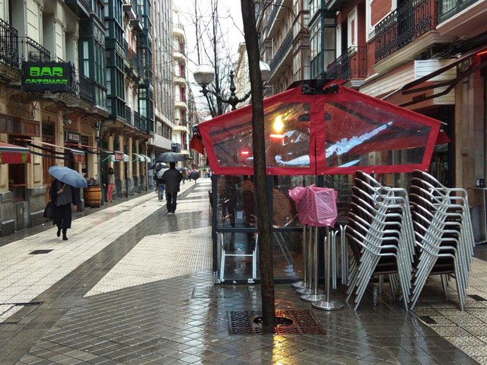 Una persona camina por Bilbao bajo la lluvia ante terrazas con el toldo recogido (archivo)