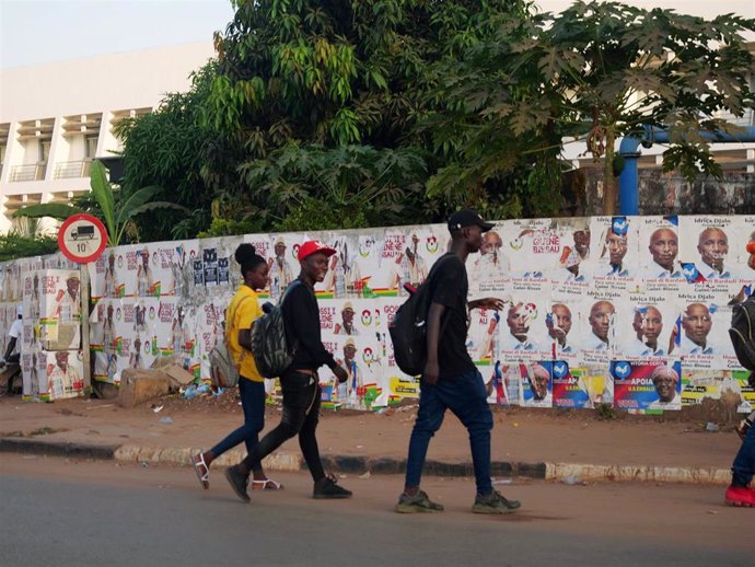 Personas caminando frente a un muro con carteles de la campaña presidencial en Bissau