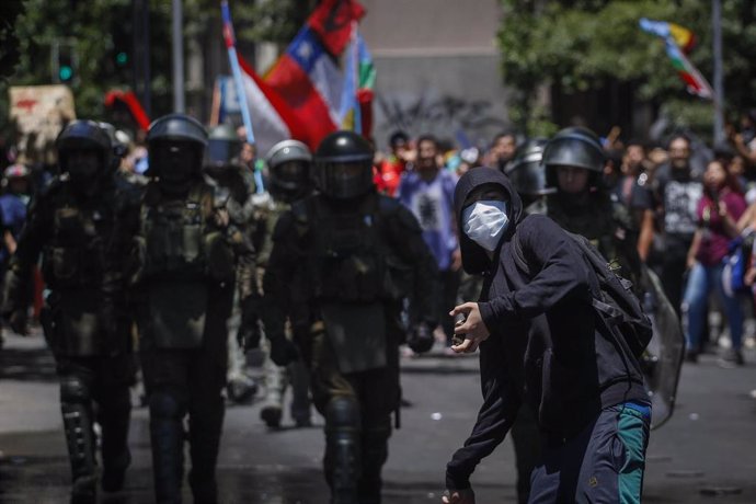 26 November 2019, Chile, Santiago: A masked demonstrator hurls stones while security forces walk towards him during a protest to demanded social reforms. Photo: Sebastian Beltran Gaete/Agencia Uno/dpa