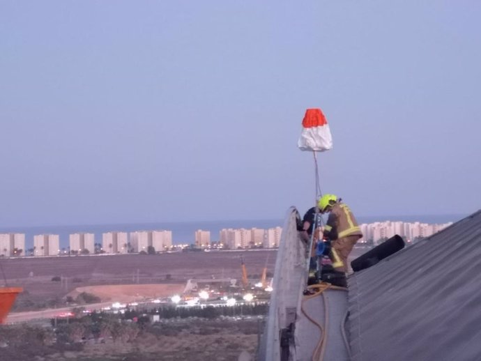 Dos bombers treballen sobre la cornisa de l'aeroport amb la platja d'Urbanova al fons.