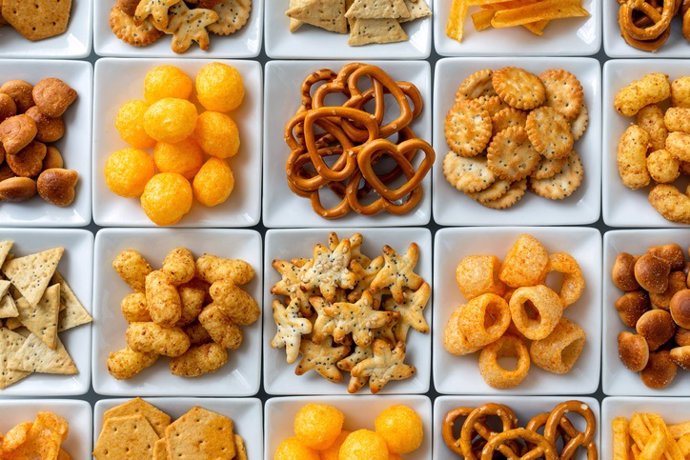 Background of many types of savory snacks in white square dishes from above.