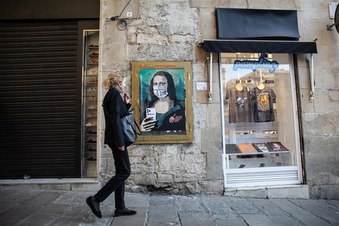 Una mujer hablando por teléfono a través de sus auriculares pasa junto a una pintura de una Gioconda con un móvil y una mascarilla, obra del grafitero TV Boy en Barcelona