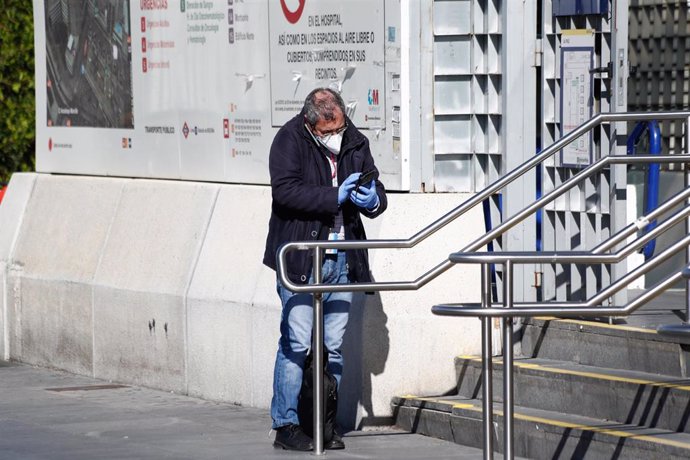 Un hombre protegido con una mascarilla y guantes consulta su móvil en la puerta de un hospital.