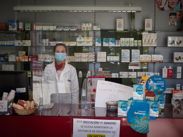 Una farmacéutica continúa trabajando protegida por una máscara sanitaria y una mampara.