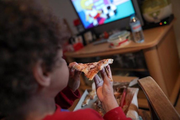 Un niño beneficiario de beca comedor en la Comunidad de Madrid come un trozo de pizza mientras ve la televisión en su casa.