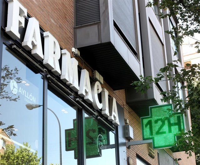 La entrada de una famacia con una señal luminosa de la cruz verde en la que pone 12h.