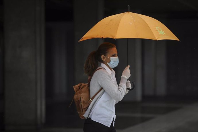 Una mujer protegida con mascarilla y guantes se resguarda de la lluvia bajo su paragüas. 