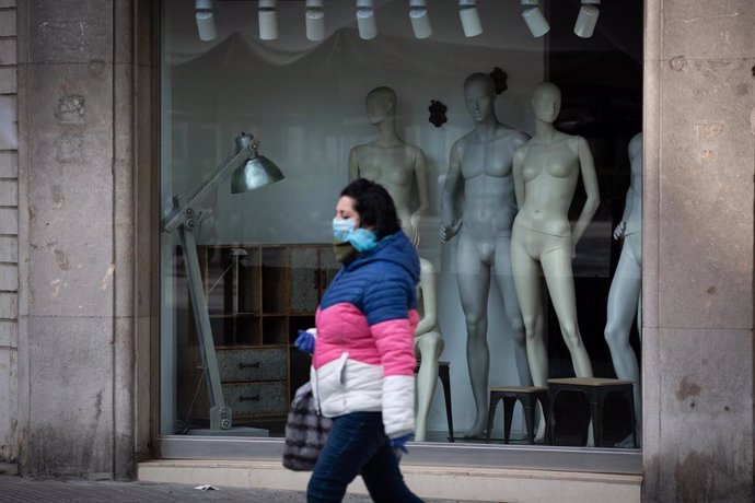 Una mujer protegida con una mascarilla pasa junto a un escaparate de una tienda cerrada en Barcelona por el estado de alarma ante el coronavirus.