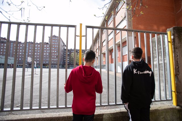 Dos adolescentes observan el patio cerrado de un colegio durante el confinamiento por el estado de alarma, cuando España es el único país que no deja a los menores salir a la calle y un día después de que la ministra de Educación anunciara que el curso es