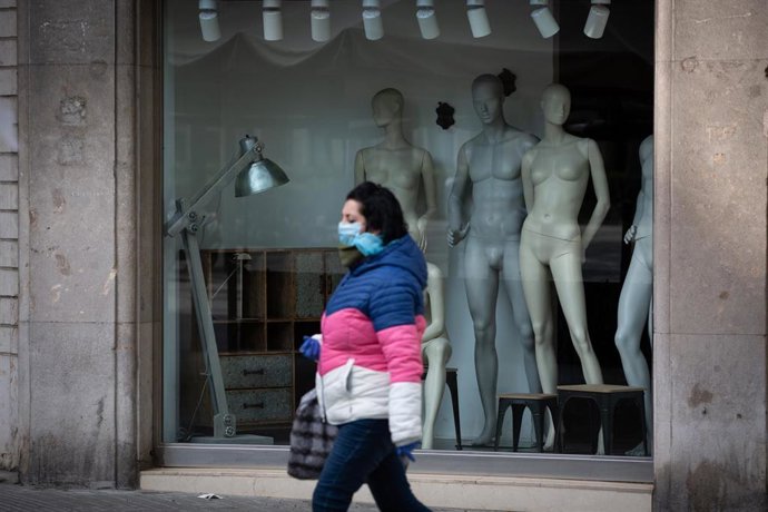 Una mujer protegida con una mascarilla pasa junto a un escaparate de una tienda cerrada en Barcelona durante el estado de alarma por el coronavirus.