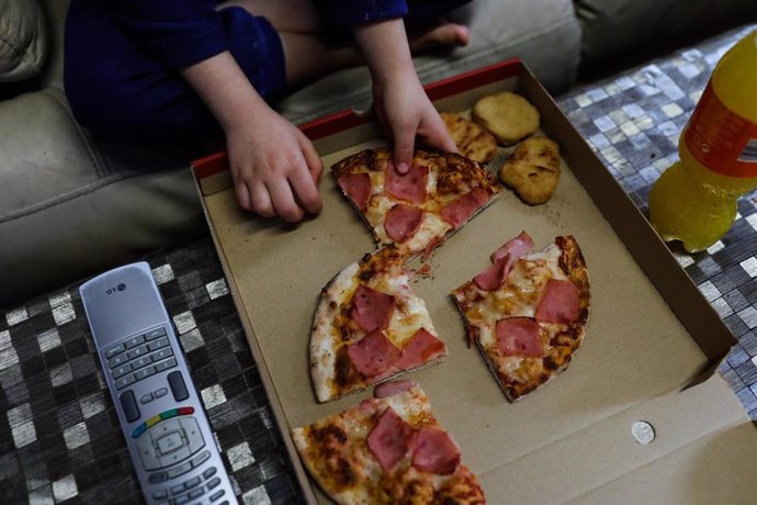 Un niño come pizza del menú infantil de Telepizza mientras ve la televisión en su casa, tras recoger el menú en un establecimiento de Telepizza, cadena que, junto a Rodilla, reparte un menú escolar a beneficiarios de beca comedor en la Comunidad de Madr