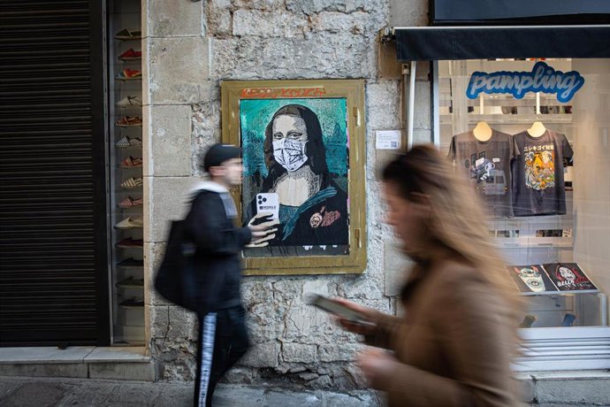 Dos personas pasan con sus móviles junto a una pintura de una Gioconda con un móvil y una mascarilla, obra del grafitero TV Boy llamada 'Mobile World Virus', en referencia a la cancelación del Mobile World Congress a causa del coronavirus, en Barcelona