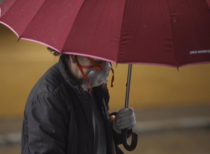 Una mujer con una peculiar mascarilla se resguarda de la lluvia bajo su paraguas, durante el día 38 del estado de alarma en el país por la crisis del coronavirus. En Sevilla (Andalucía, España), a 21 de abril de 2020.