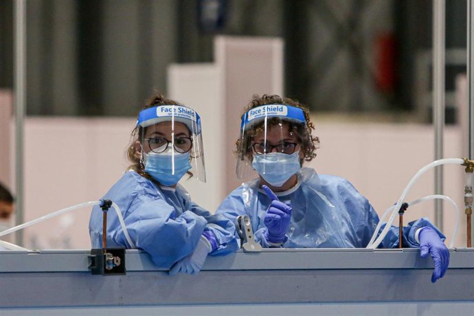Dos sanitarias protegidas con mascarilla, guantes y pantalla protectora se asoman por encima de una mampara durante la lectura de pasajes de El Quijote para los sanitarios y pacientes del Hospital temporal de IFEMA para enfermos con Covid-1.