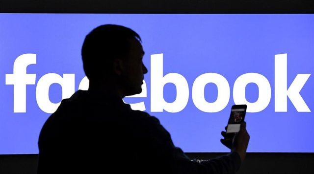 Un hombre con un teléfono móvil delante de un cartel de Facebook