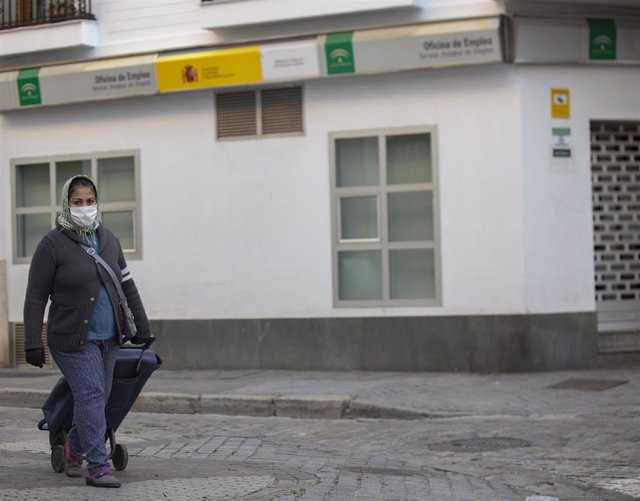 Una mujer con mascarilla portando un carro de la compra camina delante de una Oficina de Empleo.