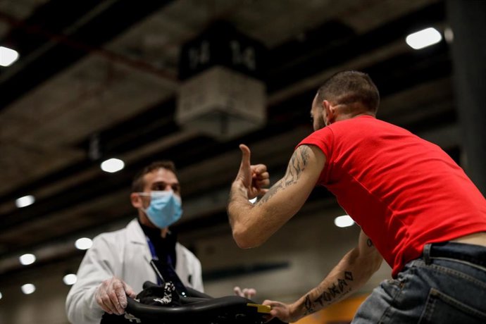 Un hombre sin hogar monta en una bicicleta estática mientras habla con un sanitario en una zona destinada al deporte en el Pabellón 14, para las personas sin hogar de IFEMA.