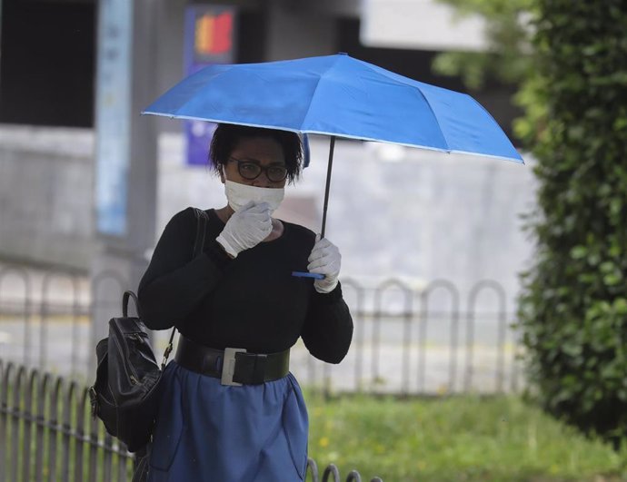 Una mujer protegida con mascarilla y guantes se resguarda de la lluvia bajo su paraguas en la quinta semana del estado de alarma por el coronavirus, Covid-19. En Sevilla (Andalucía, España), a 15 de abril 2020.