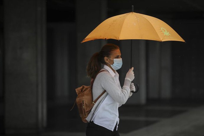 Una mujer protegida con mascarilla y guantes se resguarda de la lluvia bajo su paraguas en la quinta semana del estado de alarma por el coronavirus, Covid-19. En Sevilla (Andalucía, España), a 15 de abril 2020.