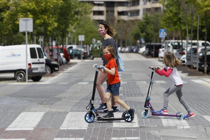 Dos niñas con mascarilla cruzan un paso de peatones montadas en patinete.