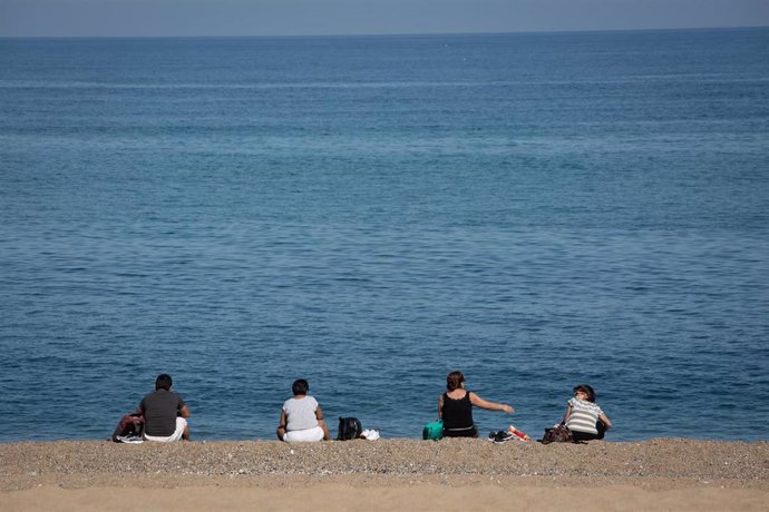 Varias personas sentadas en la Playa de la Barceloneta, en Barcelona.