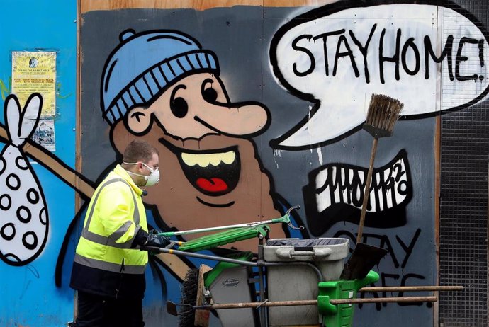 Un hombre con mascarilla junto a un mural pintado con el lema "Stay home!", "¡Quédate en casa!"