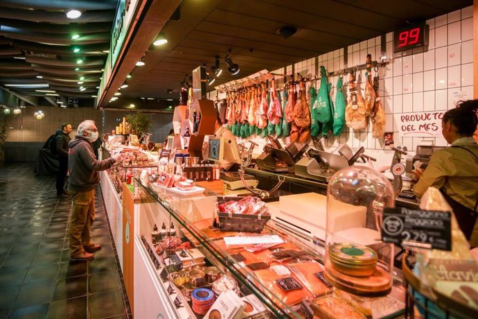 Varios clientes hacen la compra en un puesto de carne y embutidos en el mercado de San Antón, que permanece abierto y anuncia que también atiende a domicilio, en Madrid (España), a 18 de marzo de 2020.