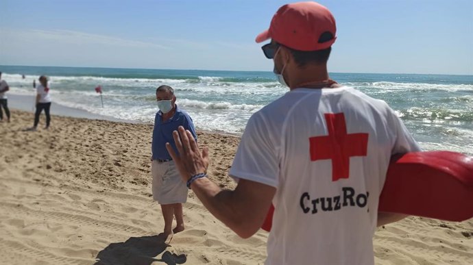 Un señor pasea por la playa con mascarilla y atiende a lo que le dice un socorrista de la Cruz Roja