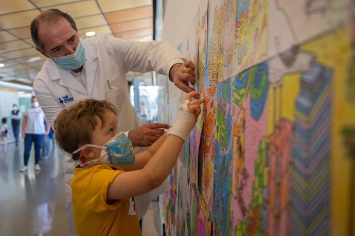 Un nen ingressat a la Vall d'Hebron pega amb l'ajuda d'un metge el seu dibuix en un mural que han installat al centre per celebrar el Dia Mundial del Nen Hospitalitzat, a Barcelona, Catalunya (Espanya), a 13 de maig de 2020.