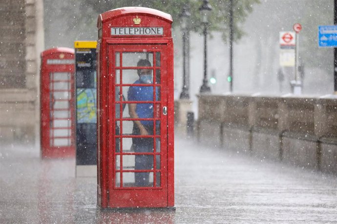 Una persona con mascarilla se refugia de la lluvia en una cabina de Londres
