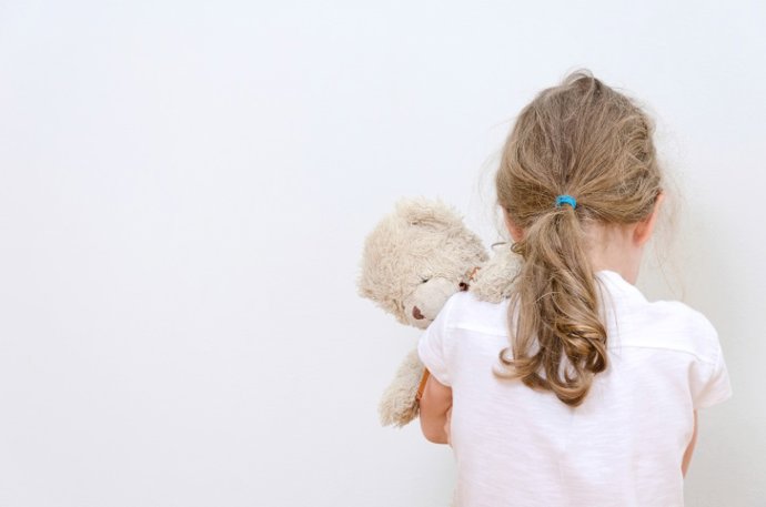 Una niña, de espaldas, con un oso de peluche.