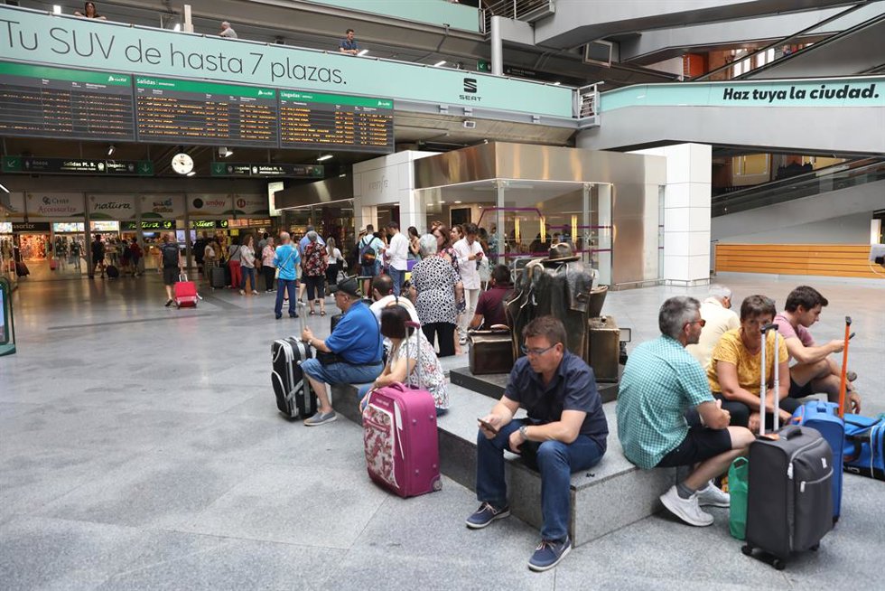 Varios pasajeros esperan sentados con sus maletas en la estación de tren Puerta de Atocha de Madrid