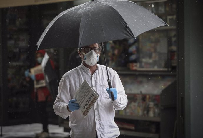 Un hombre con mascarilla y paraguas tras comprar el periódico