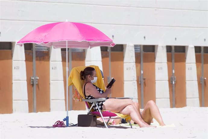Una mujer protegida con mascarilla lee en en una playa