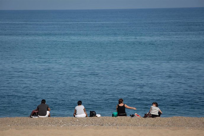 Varias personas sentadas en la Playa de la Barceloneta respetan la distancia de seguridad.
