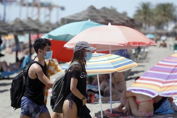 Dos personas con mascarillas mientras pasan entre sombrillas en la playa 