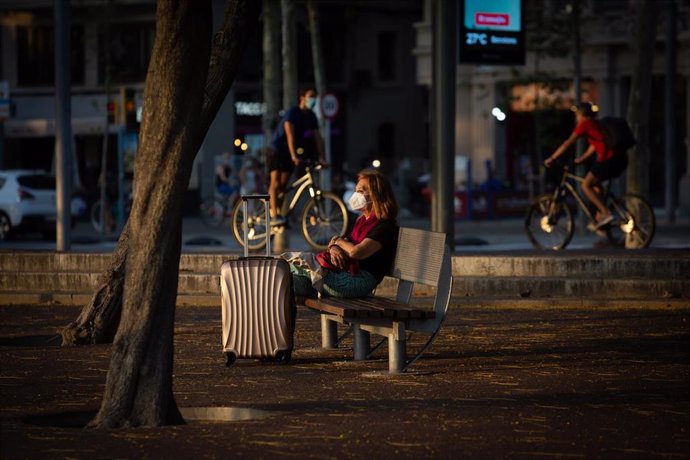 Una mujer sentada en un banco junto a una maleta, mientras varios ciclistas pasan por detrás, en Barcelona, Catalunya (España), a 28 de julio de 2020.