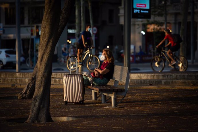 Una dona asseguda en un banc al costat d'una maleta, mentre diversos ciclistes passen per darrere, a Barcelona, Catalunya (Espanya), a 28 de juliol de 2020. 